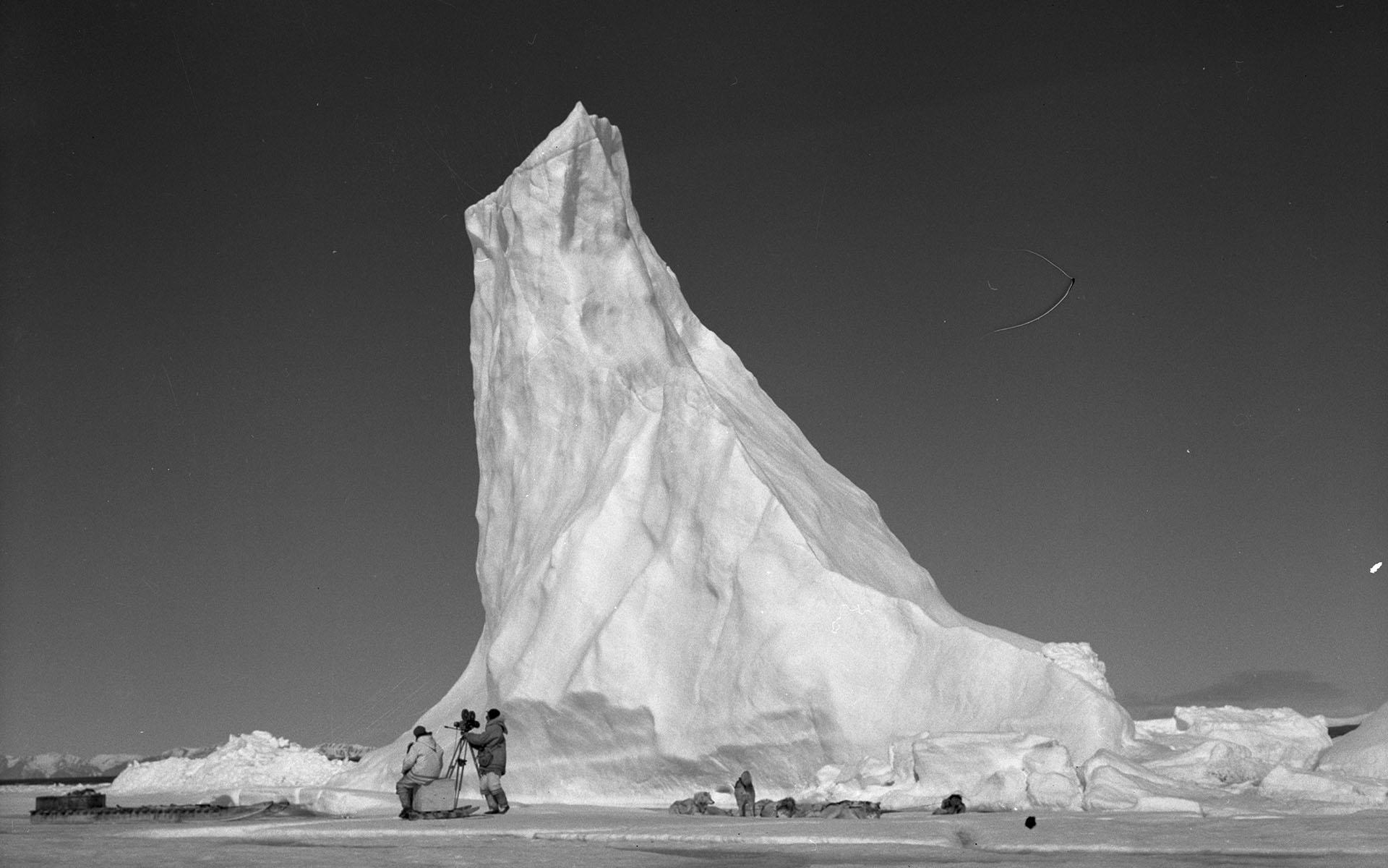 National Film Board crew setting up their camera near a towering arctic iceberg