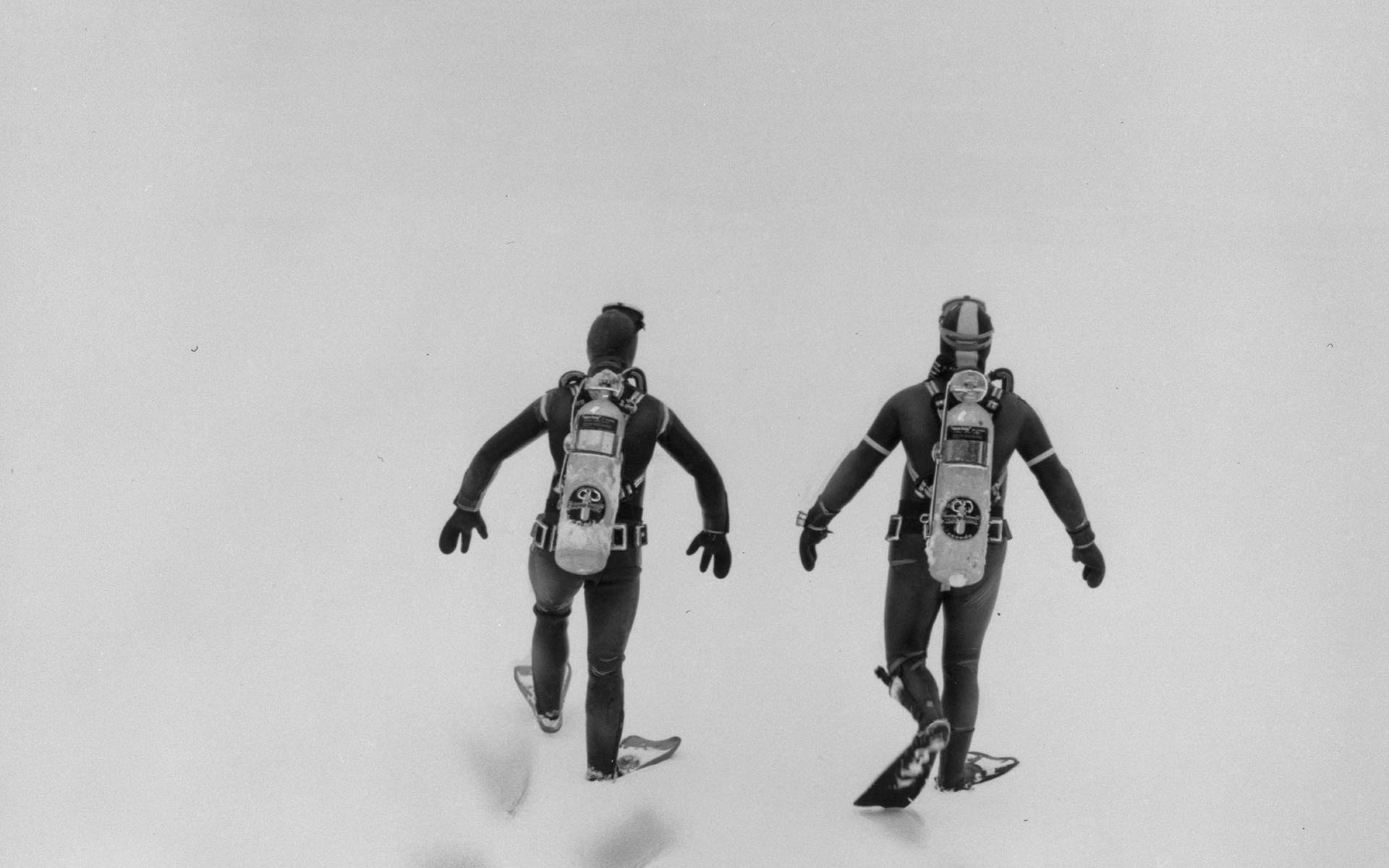 Two scuba divers, seen from behind, walking across snow